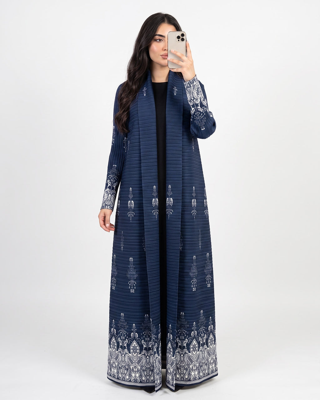 A woman takes a mirror selfie wearing a long navy blue open-front abaya with intricate white and silver floral embroidery on the sleeves and hem, set against a plain light gray background.