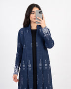 A woman takes a mirror selfie wearing a navy blue open-front cardigan with white embroidered patterns on the cuffs and hem, layered over a black top, against a plain light gray background.