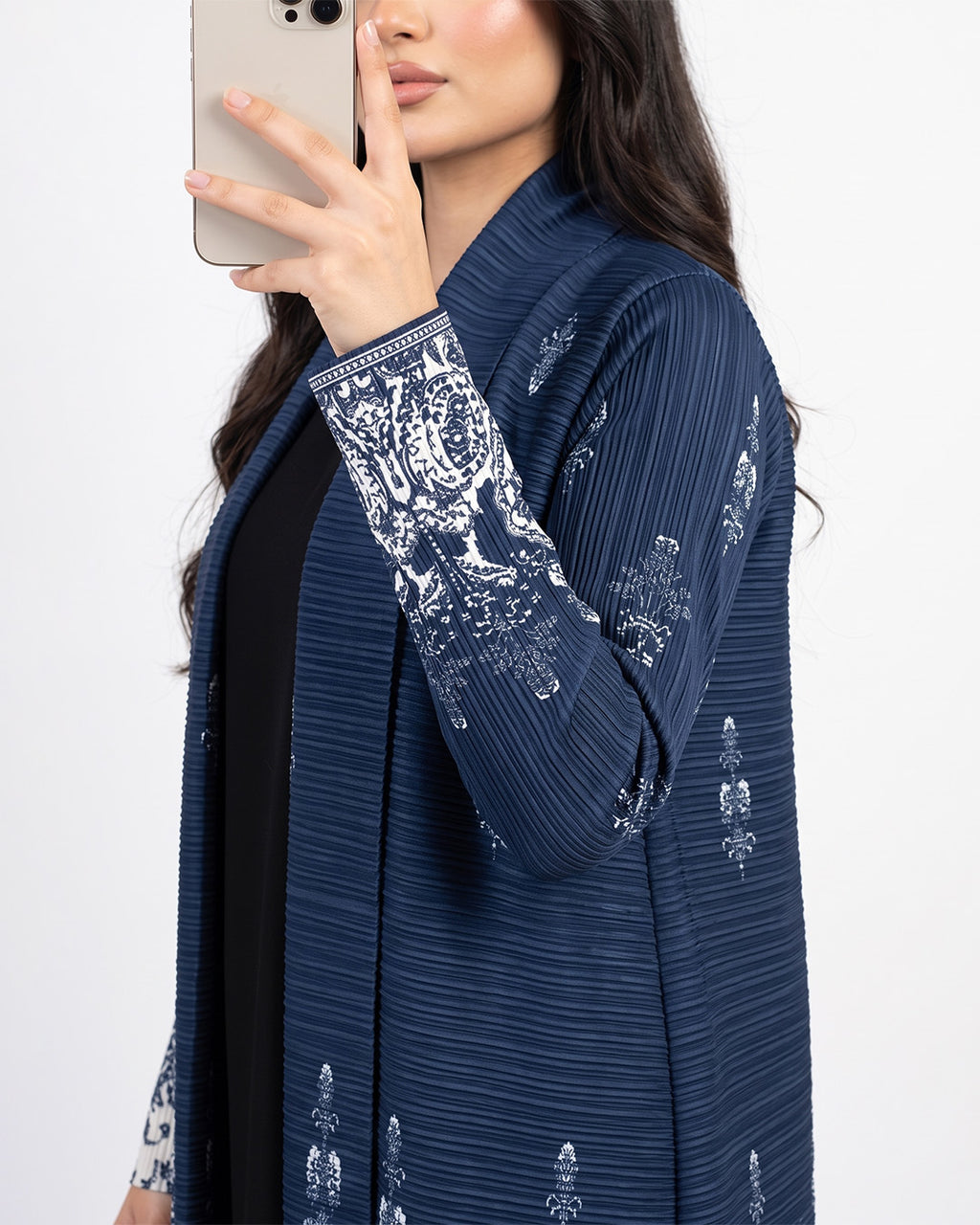 A woman takes a mirror selfie wearing a navy blue open-front cardigan with white floral embroidery on the cuffs and hem, layered over a black top against a plain white background.