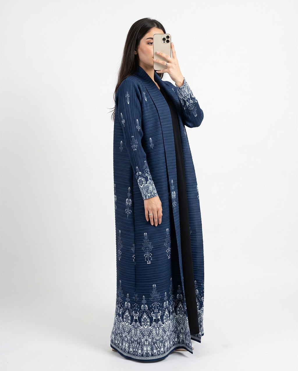 A woman takes a mirror selfie wearing a long navy blue open-front abaya with intricate white floral embroidery on the sleeves, hem, and front opening, photographed against a plain white studio background.