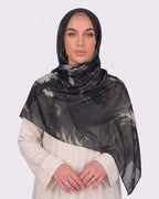 A woman wearing a moody black and smoky olive green printed modal hijab with an organic abstract tie-dye pattern, paired with a cream shirt dress.