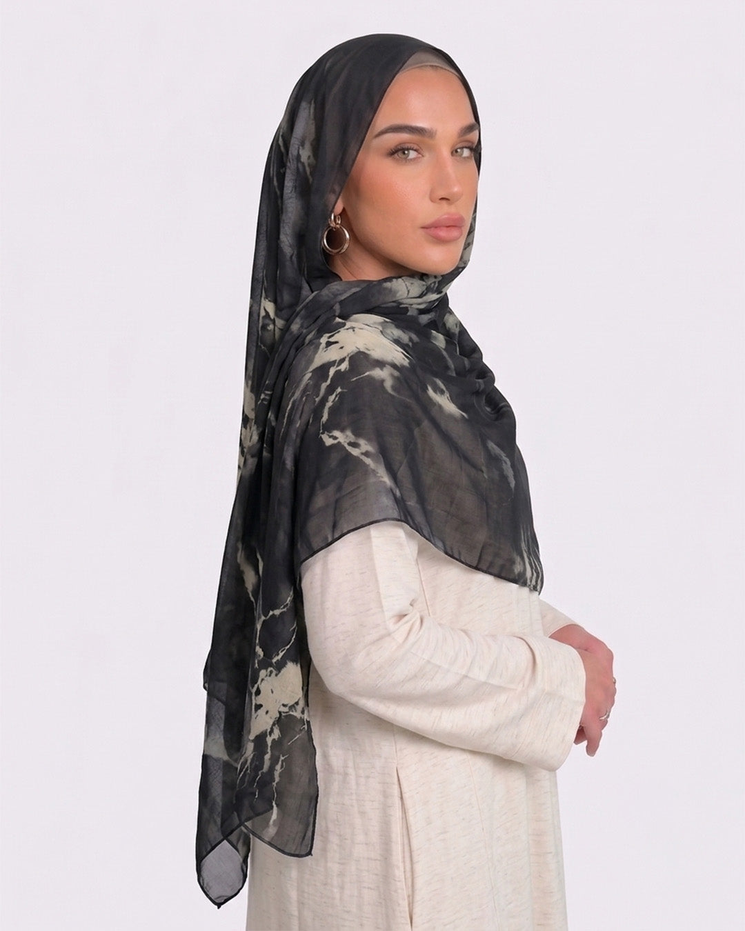 A woman wearing a moody black and smoky olive green printed modal hijab with an organic abstract tie-dye pattern, paired with a cream shirt dress.