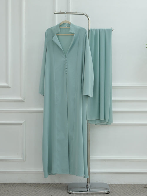 A long, loose-fitting sage green abaya dress with a buttoned front and collar, displayed on a metal hanger stand next to a matching folded garment, set against a white paneled wall.