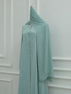A light mint green abaya dress with a matching headscarf, displayed on a mannequin against a white paneled wall.
