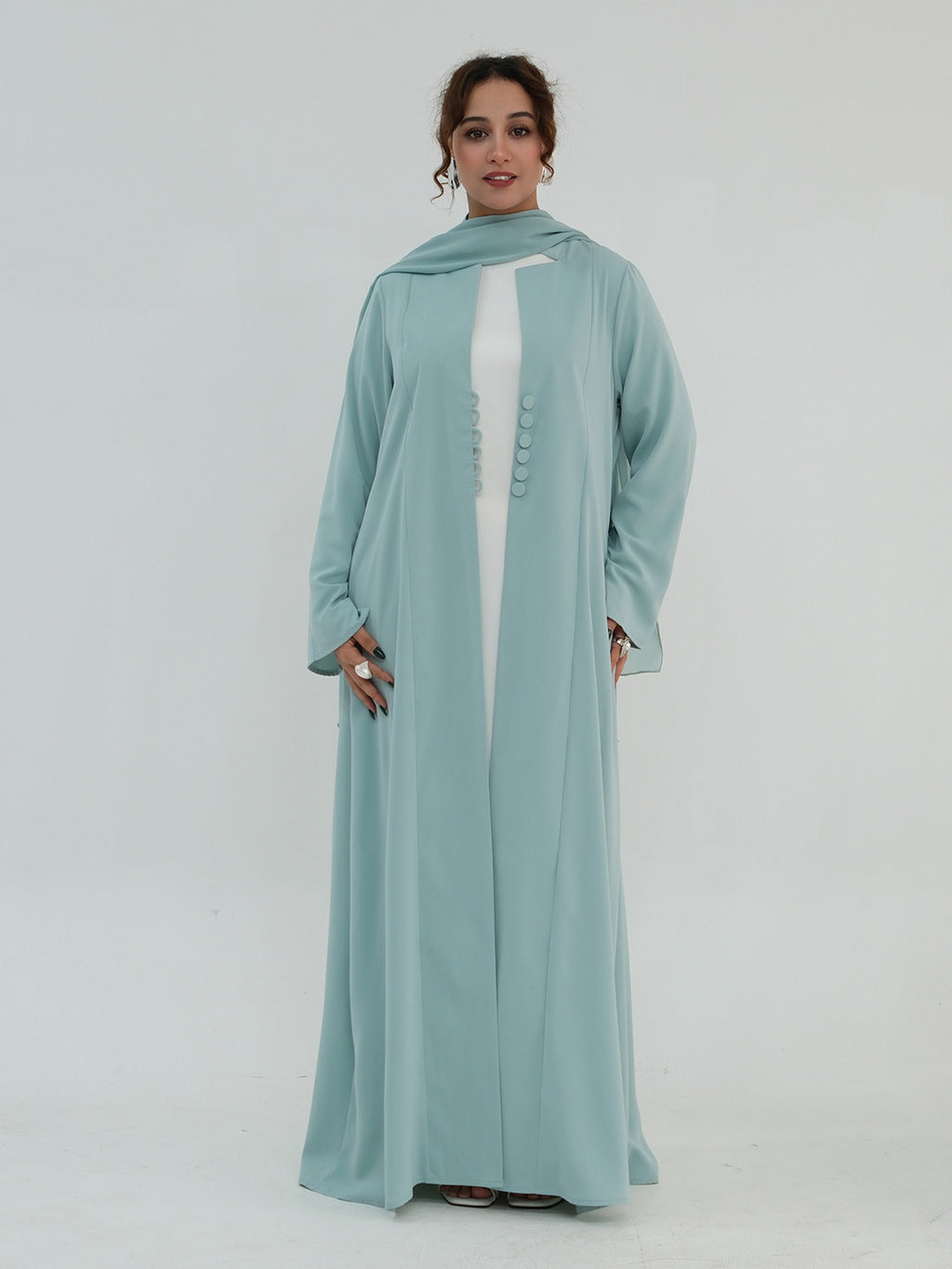 A woman modeling a long, flowing abaya in a light mint green color, worn over a white inner dress, with a matching headscarf. The abaya features a buttoned front detail down the center.