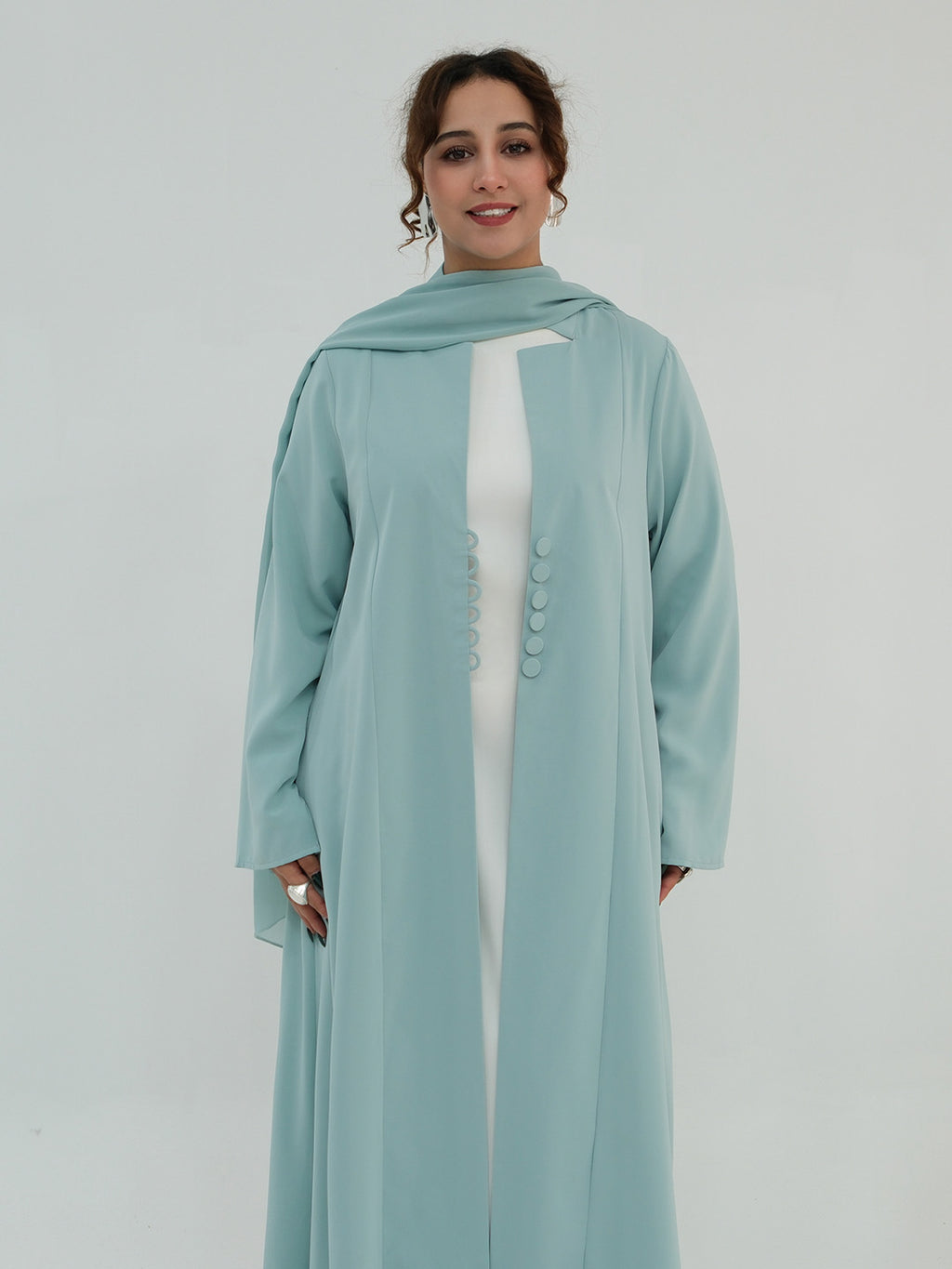 A woman modeling a long, flowing sage green abaya with a matching headscarf, worn over a white inner dress. The abaya features a high neckline, wide sleeves, and a decorative row of matching buttons down the front center.