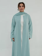 A woman modeling a long, flowing sage green abaya with a matching headscarf, worn over a white inner dress. The abaya features a high neckline, wide sleeves, and a decorative row of matching buttons down the front center.