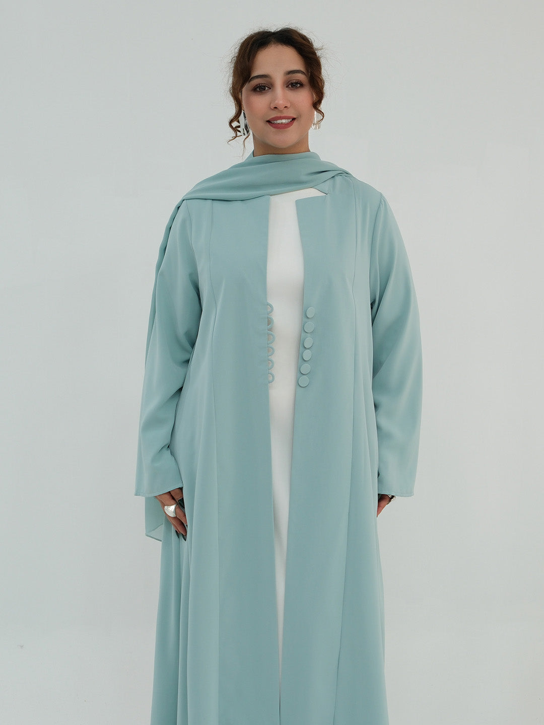 A woman modeling a long, flowing sage green abaya with a matching headscarf, worn over a white inner dress. The abaya features a high neckline, wide sleeves, and a decorative row of matching buttons down the front center.