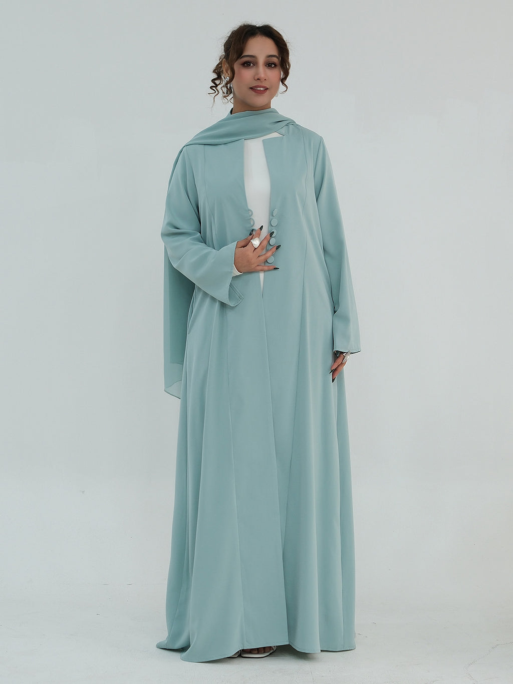 A woman modeling a long flowing abaya dress in a dusty light blue-green color, featuring a matching headscarf and a white inner lining visible at the chest, set against a plain light gray studio background.