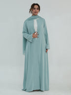 A woman modeling a long flowing abaya dress in a dusty light blue-green color, featuring a matching headscarf and a white inner lining visible at the chest, set against a plain light gray studio background.