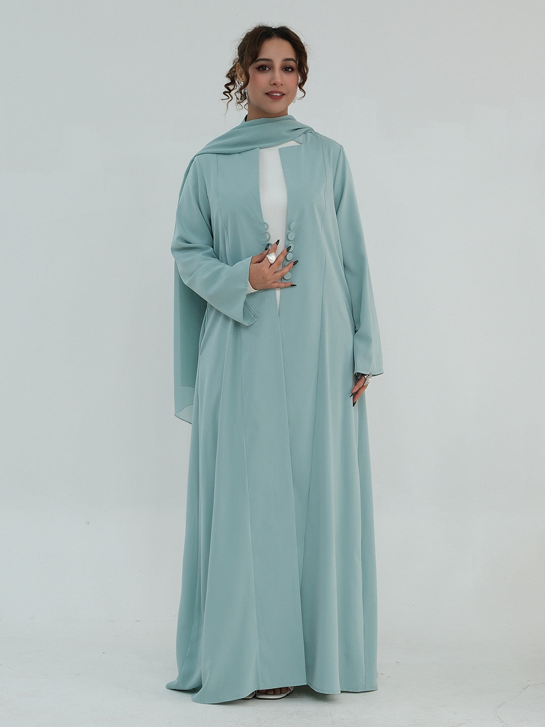A woman modeling a long flowing abaya dress in a dusty light blue-green color, featuring a matching headscarf and a white inner lining visible at the chest, set against a plain light gray studio background.