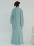 A woman viewed from behind wearing a flowing, long-sleeved abaya in a soft dusty blue-green color against a plain white studio background.