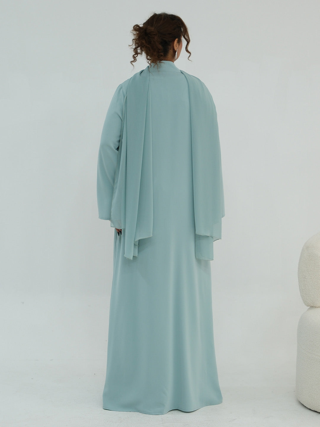 A woman viewed from behind wearing a flowing, long-sleeved abaya in a soft dusty blue-green color against a plain white studio background.