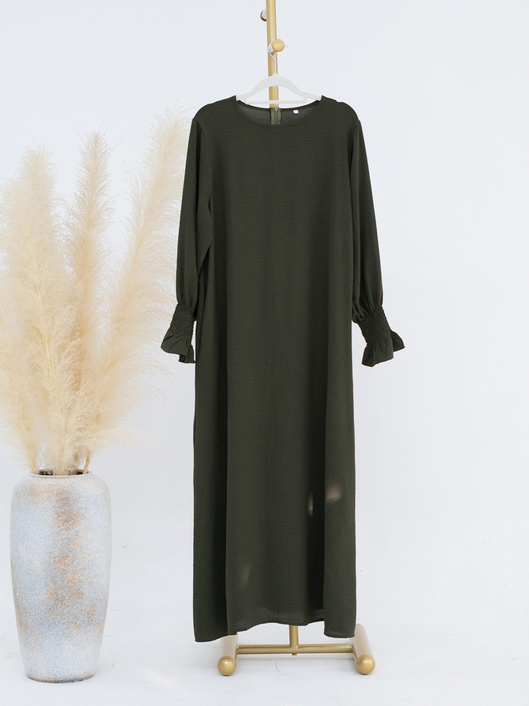 An olive green long-sleeve maxi dress displayed on a gold hanger stand, next to a decorative vase with dried pampas grass against a plain white background.