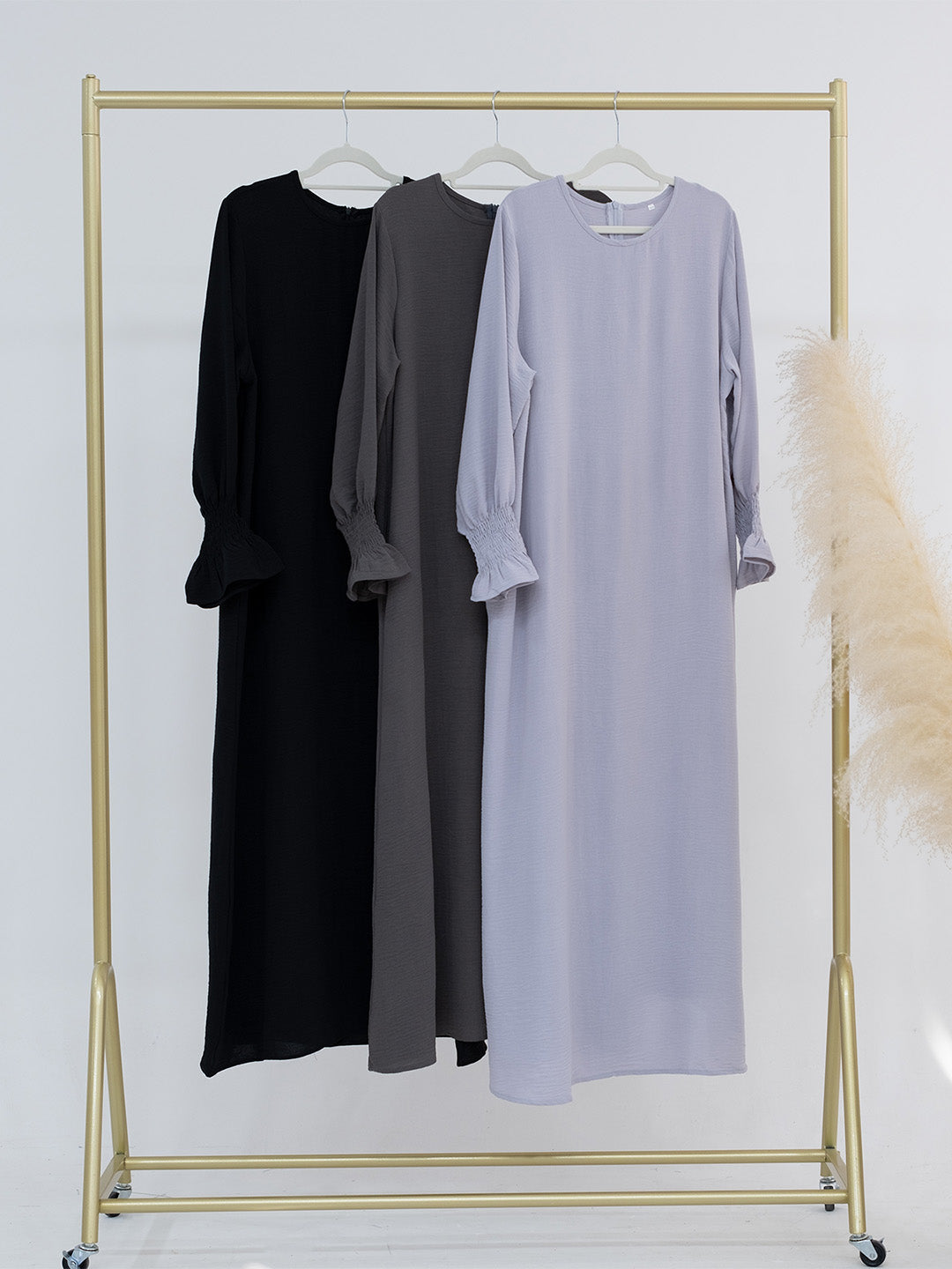 Three long-sleeve midi dresses hanging on a gold clothing rack against a white background: black, dark gray, and light lavender. A beige pampas grass decoration is visible on the right side.
