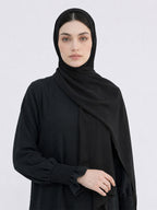 A woman wearing a black hijab and a black long-sleeve abaya with gathered cuffs, photographed against a light gray studio background.