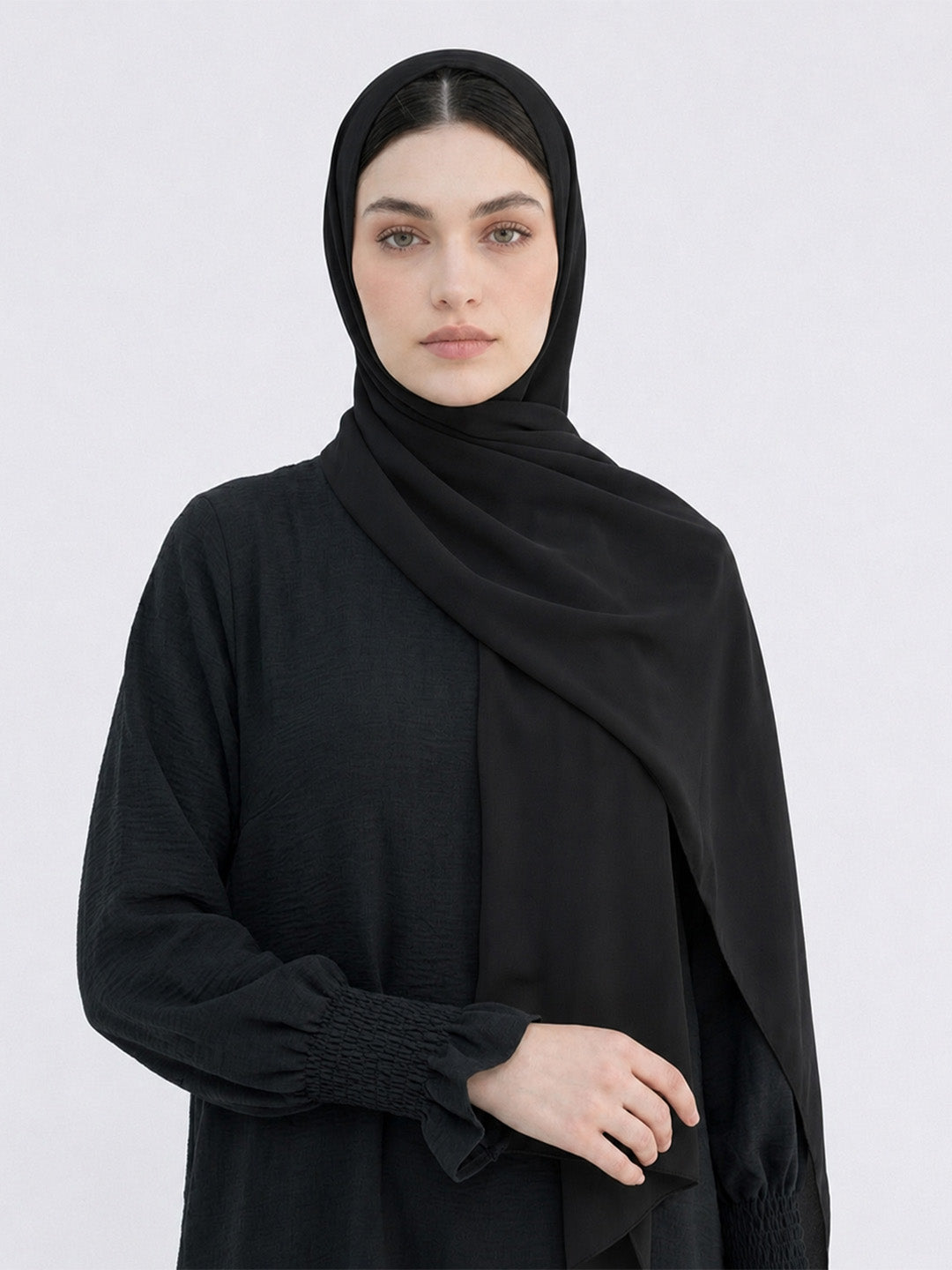 A woman wearing a black hijab and a black long-sleeve abaya with gathered cuffs, photographed against a light gray studio background.
