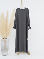 A dark gray long-sleeve maxi dress with ruffled cuffs, displayed on a gold hanger stand next to a decorative vase with dried pampas grass against a white background.