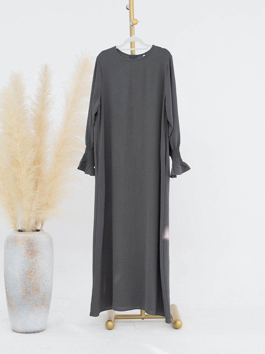 A dark gray long-sleeve maxi dress with ruffled cuffs, displayed on a gold hanger stand next to a decorative vase with dried pampas grass against a white background.