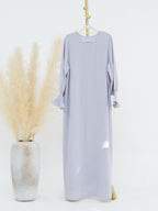A light lavender maxi dress with three-quarter ruffled sleeves hangs on a hanger against a white studio background, styled next to a textured vase with dried pampas grass.