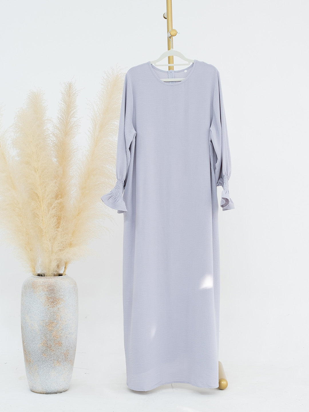 A light lavender maxi dress with three-quarter ruffled sleeves hangs on a hanger against a white studio background, styled next to a textured vase with dried pampas grass.