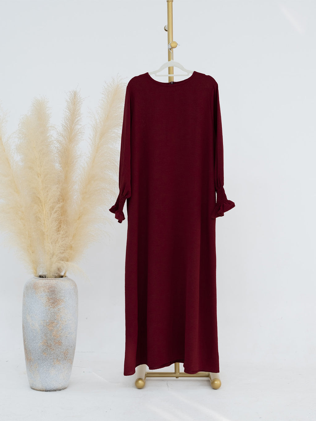 A long-sleeve maxi dress in deep burgundy is displayed on a gold hanger stand next to a tall textured vase filled with pampas grass against a clean white background.