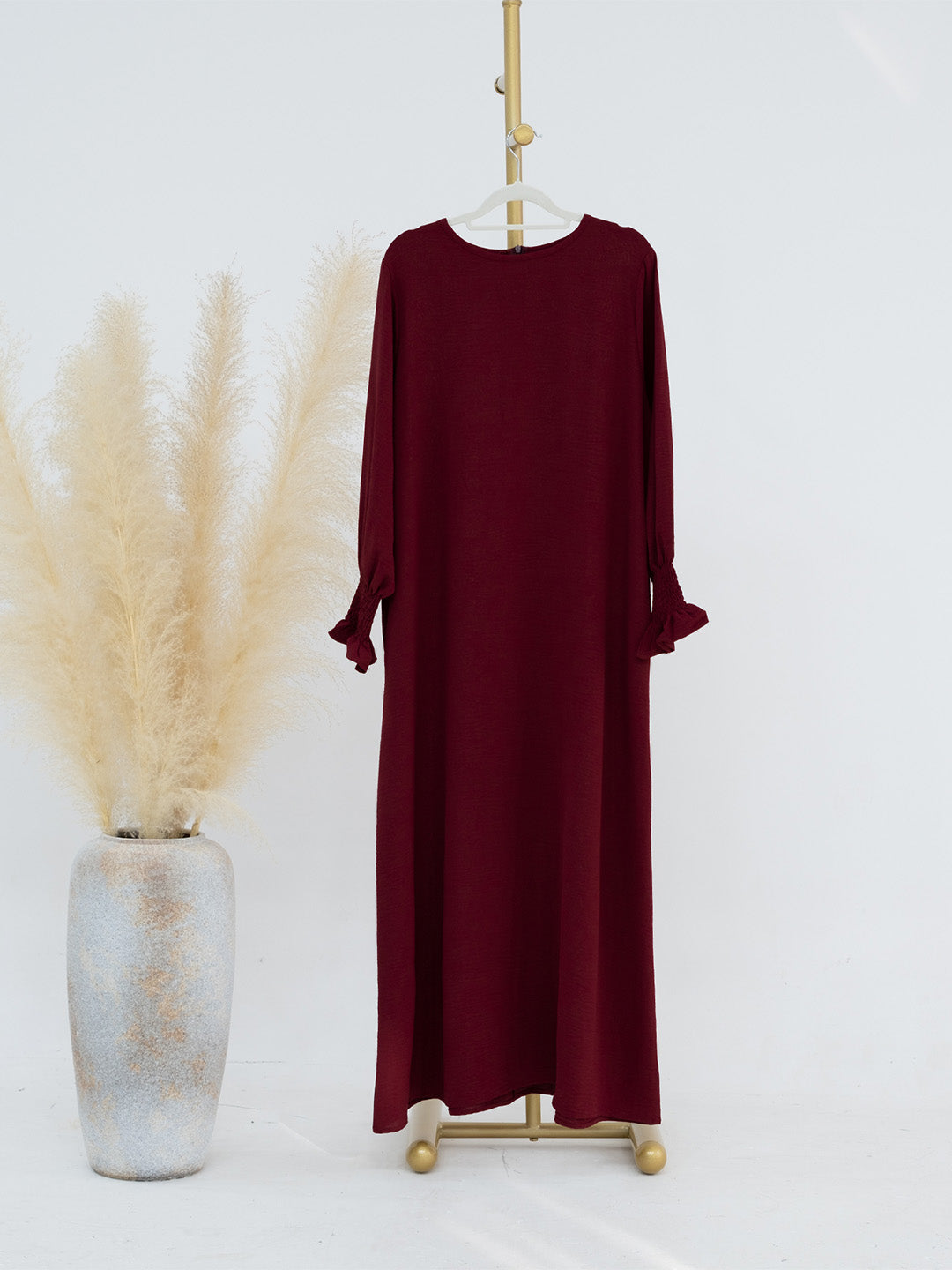 A long-sleeve maxi dress in deep burgundy is displayed on a gold hanger stand next to a tall textured vase filled with pampas grass against a clean white background.
