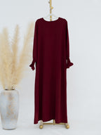 A long-sleeve maxi dress in deep burgundy, displayed on a gold dress form stand next to a decorative vase with pampas grass against a plain white background.