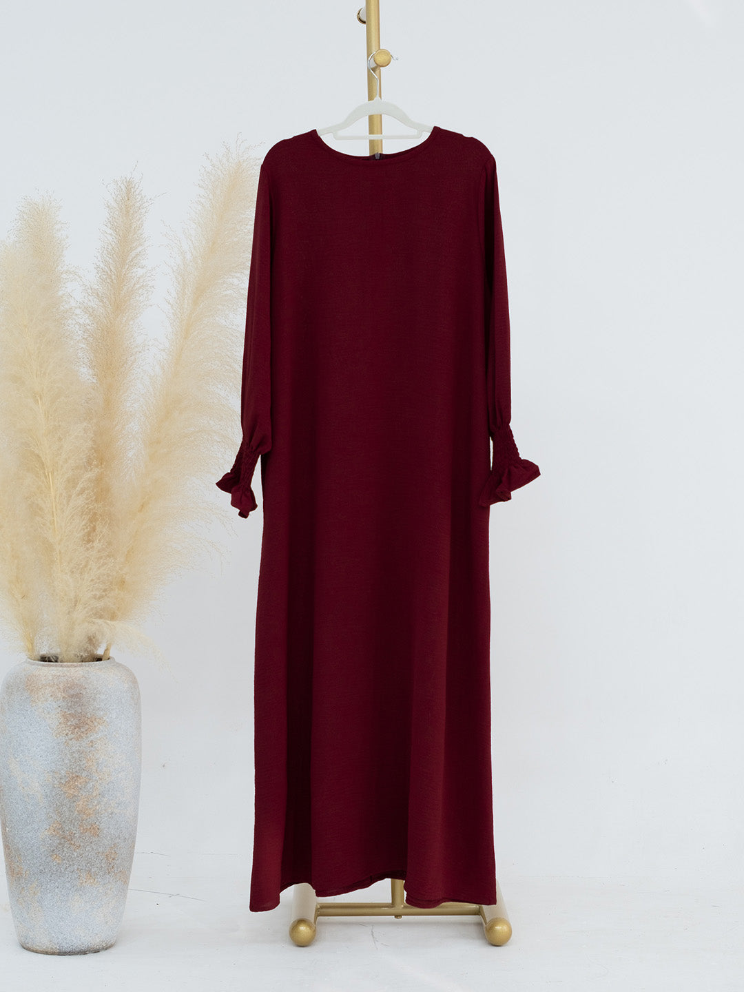A long-sleeve maxi dress in deep burgundy, displayed on a gold dress form stand next to a decorative vase with pampas grass against a plain white background.