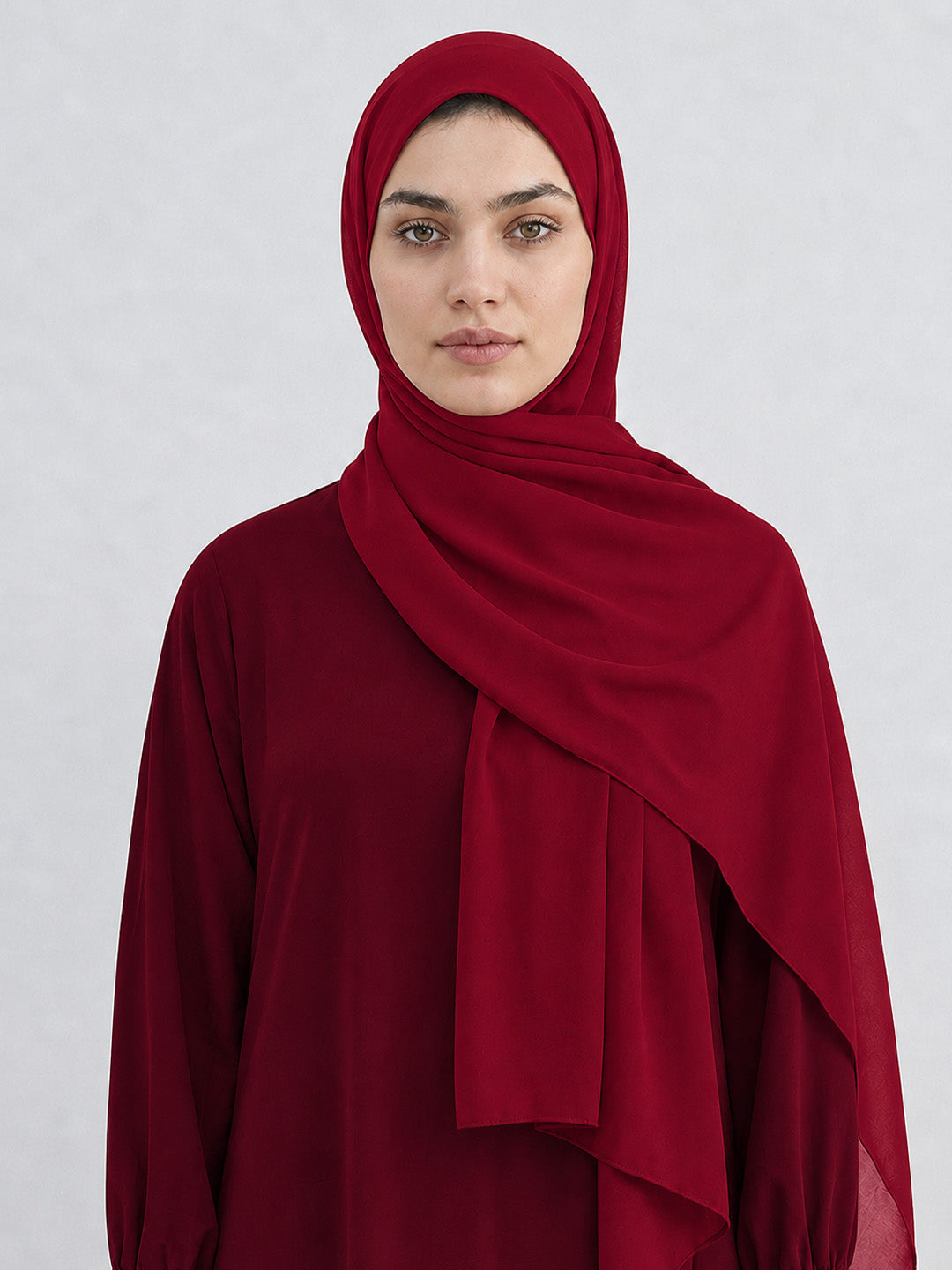 A woman wearing a deep red hijab and matching long-sleeve top, photographed against a light gray studio background.