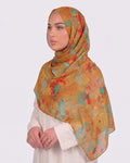 A woman wearing a vibrant mustard ochre printed modal hijab featuring abstract watercolor splatters of turquoise blue and fiery red, paired with a cream outfit.