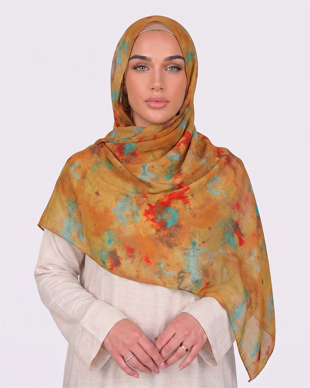 A woman wearing a vibrant mustard ochre printed modal hijab featuring abstract watercolor splatters of turquoise blue and fiery red, paired with a cream outfit.