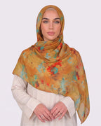 A woman wearing a vibrant mustard ochre printed modal hijab featuring abstract watercolor splatters of turquoise blue and fiery red, paired with a cream outfit.