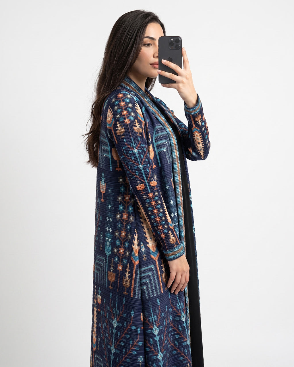 A woman takes a mirror selfie wearing a long-sleeved navy blue open-front cardigan with a detailed geometric and tribal pattern in orange, beige, and teal, set against a plain light gray background.