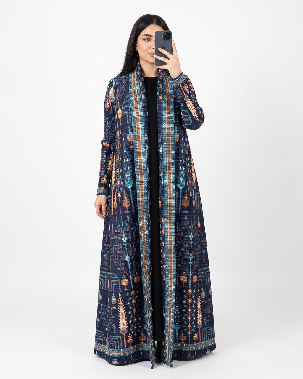 A woman takes a mirror selfie wearing a long navy blue open-front kaftan dress with an intricate multicolor geometric and tribal pattern in blue, orange, and beige, over a black top, against a plain white studio background.