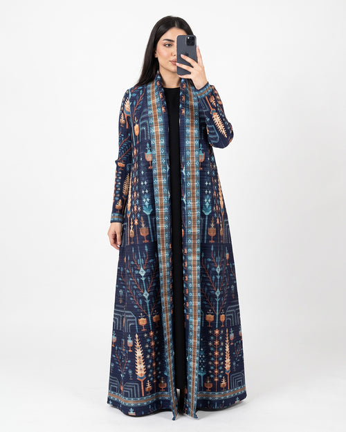 A woman takes a mirror selfie wearing a long navy blue open-front kaftan dress with an intricate multicolor geometric and tribal pattern in blue, orange, and beige, over a black top, against a plain white studio background.