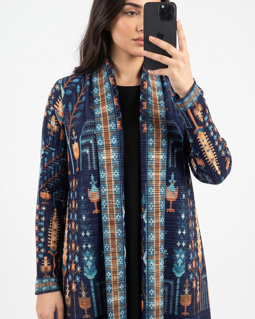 A woman takes a mirror selfie wearing a long-sleeve open-front cardigan with a complex geometric and tribal pattern in navy blue, teal, orange, and brown over a black top, set against a plain light gray background.