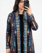 A woman takes a mirror selfie wearing a long-sleeve open-front cardigan with a complex geometric and tribal pattern in navy blue, teal, orange, and brown over a black top, set against a plain light gray background.