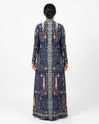 A woman viewed from the back wearing a long-sleeved navy blue maxi dress with an intricate geometric and tribal pattern in beige, orange, and light blue, set against a plain white studio background.
