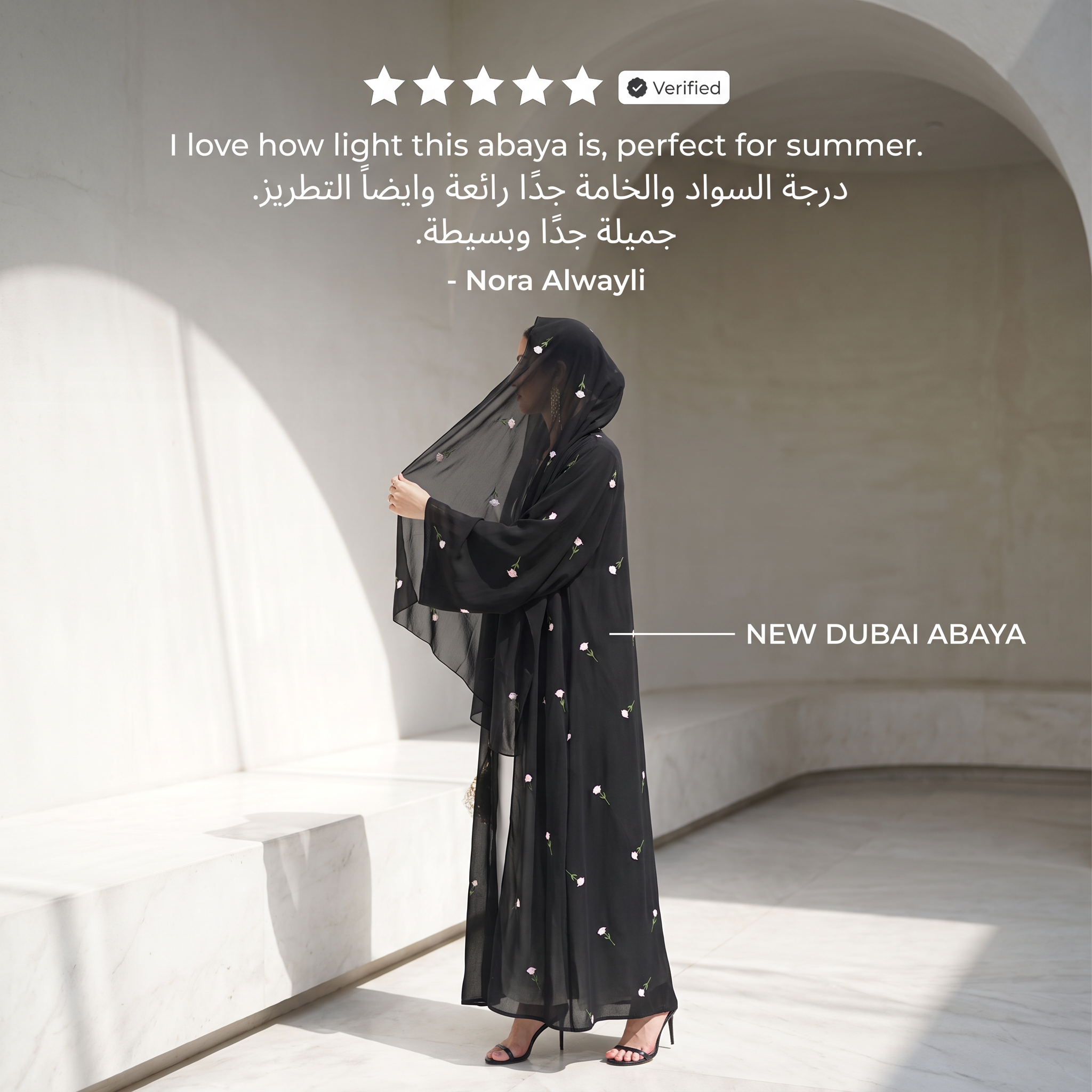 A woman wearing a black abaya with small white embroidered star-like details stands in a sunlit architectural setting with white walls and arches. The abaya has a sheer hood and wide sleeves.