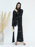 A woman modeling a black long-sleeved modest outer garment with small white floral embroidery, worn over a white dress and black hijab, standing against a light gray studio background with a pampas grass vase on the left.