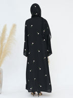A woman viewed from the back wearing a black modest dress with a matching headscarf, decorated with small white floral prints. She is standing against a plain white studio background with dried pampas grass on the left.