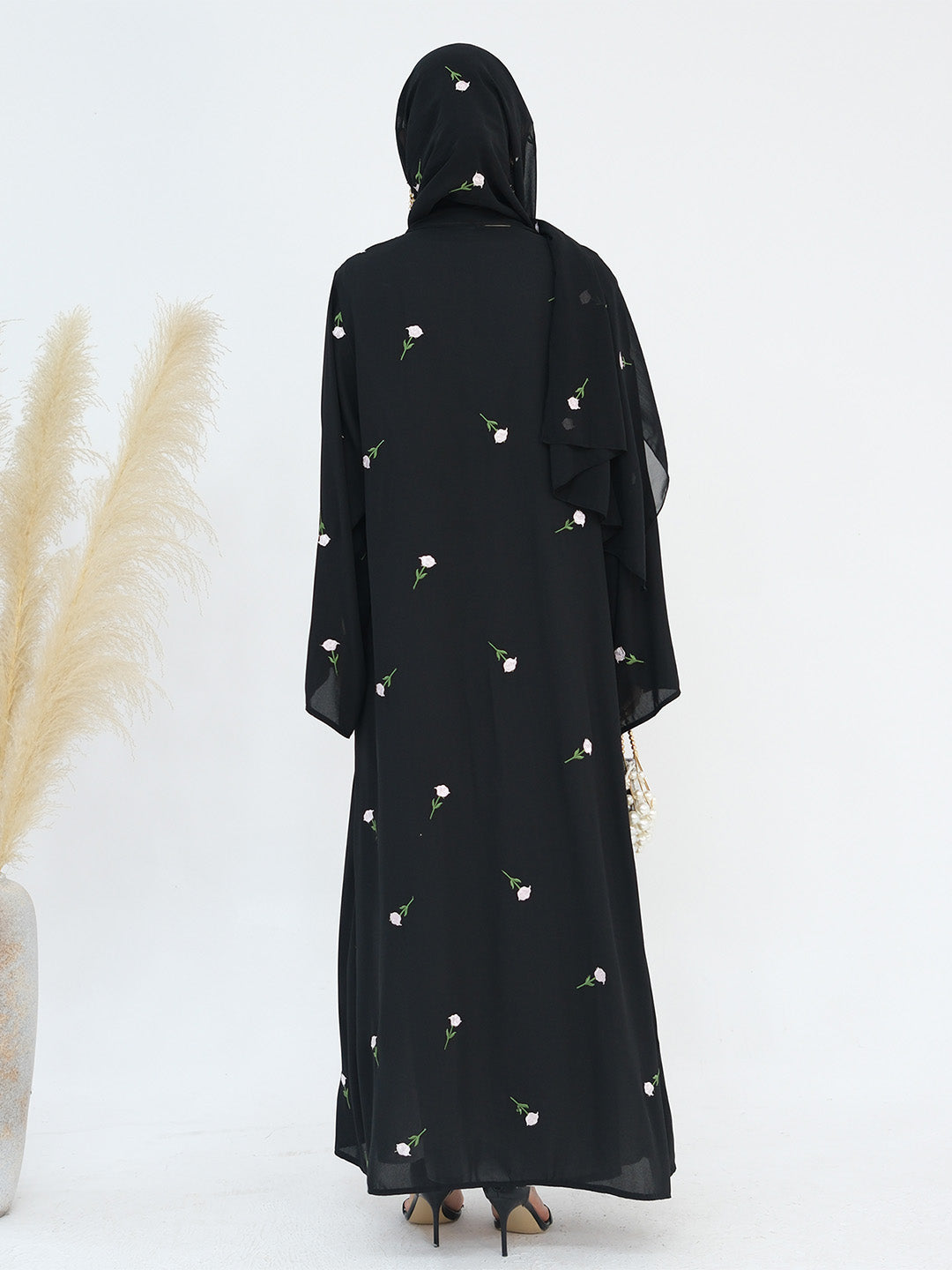A woman viewed from the back wearing a black modest dress with a matching headscarf, decorated with small white floral prints. She is standing against a plain white studio background with dried pampas grass on the left.