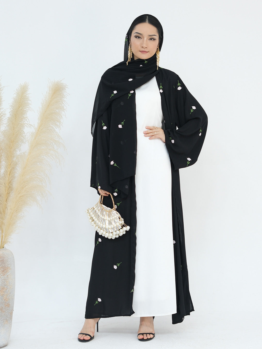 A woman modeling a black long abaya with delicate white floral embroidery, worn over a white maxi dress, a black hijab, and holding a pearl-patterned handbag.