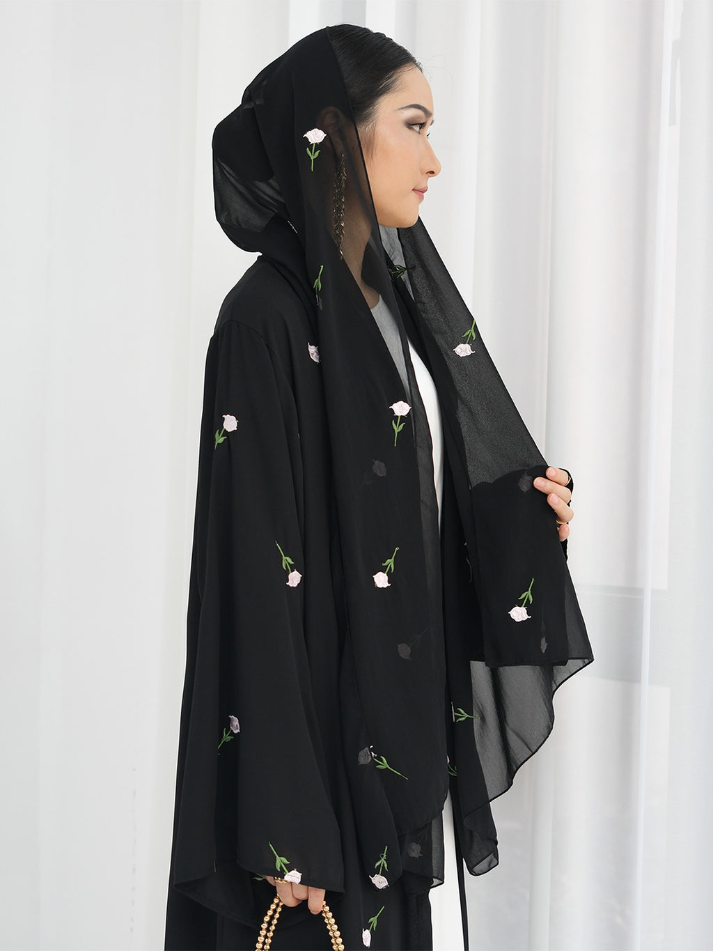 A woman wearing a black abaya with delicate pink and green floral embroidery, paired with a matching black hijab, standing against a white curtain background.