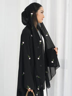 A woman wearing a black abaya with delicate pink and green floral embroidery, paired with a matching black hijab, standing against a white curtain background.