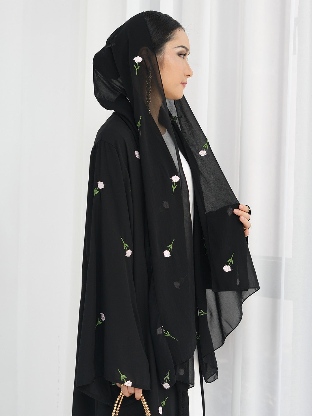 A woman wearing a black abaya with delicate pink and green floral embroidery, paired with a matching black hijab, standing against a white curtain background.