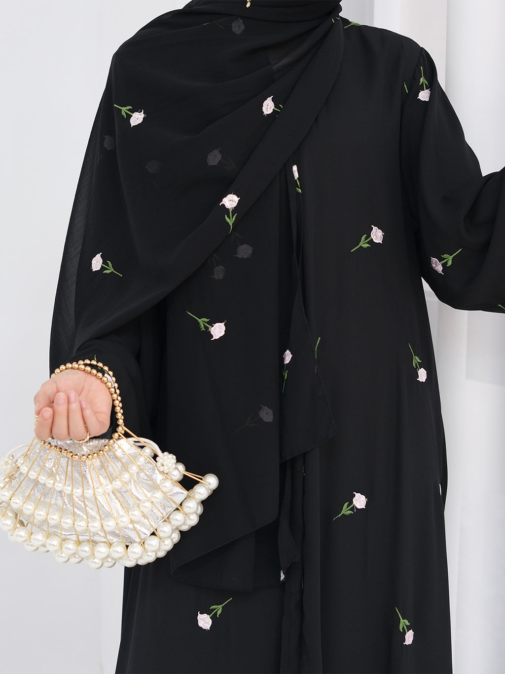 A person wearing a black modest outfit with a matching draped scarf or abaya decorated with small pink and green floral prints, holding a pearl-beaded handbag with a gold chain.