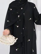 A person wearing a black modest outfit with a matching draped scarf or abaya decorated with small pink and green floral prints, holding a pearl-beaded handbag with a gold chain.