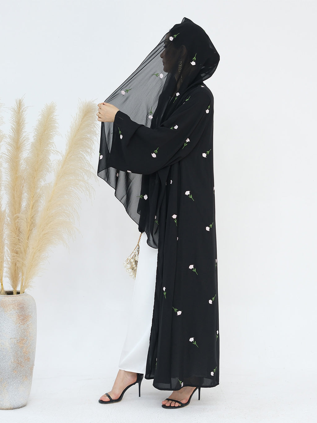 A woman models a black hooded abaya with delicate white floral embroidery, paired with white pants and black heels, standing next to a vase of pampas grass against a white studio background.